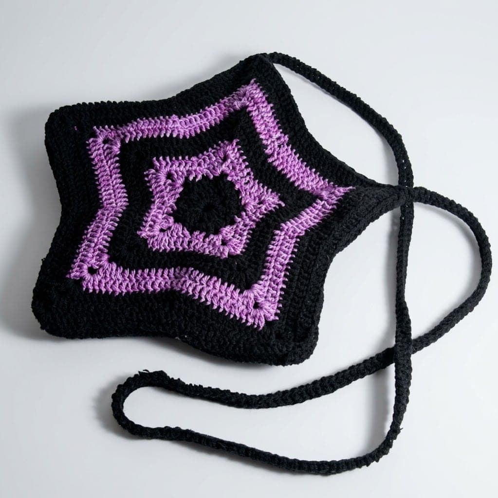 Com base preta e detalhes geométricos em roxo, ela possui um visual marcante e versátil, perfeito para quem gosta de peças artesanais com personalidade. A bolsa conta com alça longa, permitindo que se